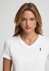 T-shirt bianca in cotone con scollo a V, caratterizzata da un piccolo logo blu navy raffigurante un golfista sul petto a sinistra. Design semplice e classico con maniche corte.