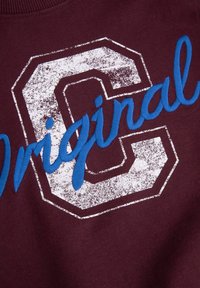 Bordowa bluza z teksturowanym białym nadrukiem litery "C" oraz niebieskim haftowanym napisem "Original". Wykonana z miękkiego materiału bawełnianego z ribbowanym dekoltem.