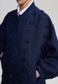 Navy blue bomber jacket featuring textured floral embroidery, snap buttons, ribbed collar, and side pockets. Fabric has a slightly soft finish.