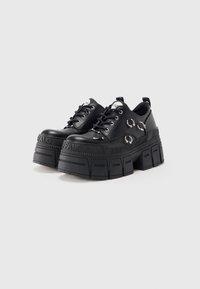 Sapatos plataforma pretos com solas grossas, argolas de metal prateado e tachas nas laterais, na frente e nos atacadores, exibidos sobre um fundo branco.