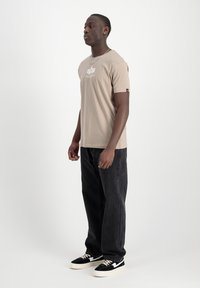 Bežs īsām piedurknēm T-krekls ar baltu logotipu, saskaņots ar tumšu džinsu biksēm un melniem sporta apaviem ar baltām akcentiem. Vienkāršs un ikdienišķs dizains.