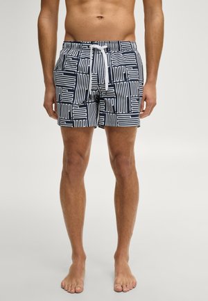 Mann trägt marineblaue und weiße Badehose mit geometrischem Muster und weißem Kordelzug, steht barfuß vor schlichtem Hintergrund.