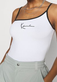 Biały top na ramiączkach z cienkimi czarnymi paskami, z czarnym logo sygnatury z przodu. Noszony z jasnoszarymi spodniami o wysokim stanie.