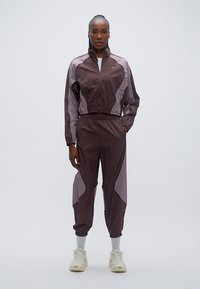 Βυσσινί αθλητική φόρμα με ανοιχτό μωβ λεπτομέρειες, που περιλαμβάνει μπουφάν με φερμουάρ και παντελόνι με ελαστική μέση. Κατασκευασμένη από ελαφρύ, λείο υλικό.