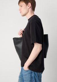 Svart lær totebag med et minimalistisk design, med doble håndtak og en strukturert form, bæres av en person i svart skjorte og jeans.