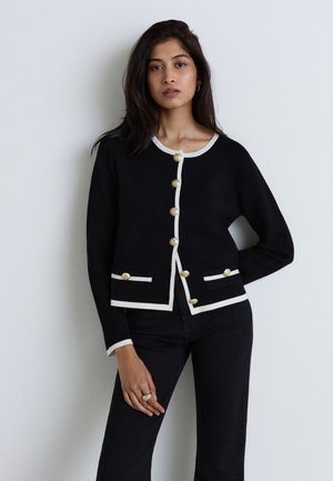 Femme aux longs cheveux foncés porte un cardigan noir avec une bordure blanche et des boutons dorés, associé à un pantalon noir, debout devant un mur clair uni.