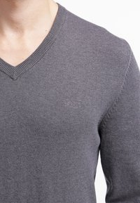 Szary sweter z dzianiny z dekoltem w serek, o miękkiej fakturze i subtelnym logo marki na piersi. Posiada prążkowany dekolt i mankiety.