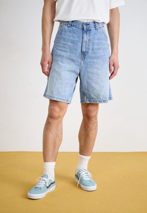 Homme portant un short en jean bleu clair, des chaussettes blanches montantes et des baskets bleu clair, debout sur un sol moutarde avec un fond blanc.