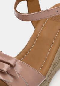 Ροζ σανδάλι από πατέντα δέρμα με διακοσμητικό φιόγκο, πλεκτή σόλα εσπαντρίγιας και καφέ υφασμάτινη βάση πέλματος, με λευκές ραφές.