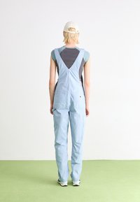 Helleblaue Denim-Latzhosen mit V-förmigem Rücken, verstellbaren Trägern und mehreren Taschen; getragen über einem dunkelgrauen Shirt.