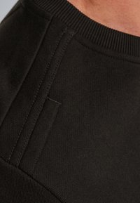 Primer plano de un sudadera negra, con tejido texturizado, cuello acanalado y costuras visibles a lo largo de las costuras.
