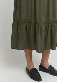 Vestito verde oliva fino alla caviglia con tessuto texturizzato e orlo arricciato. Abbinato a scarpe slip-on in pelle nera. Design semplice senza motivi.