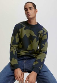 Grüner und marineblauer, gemusterter Strickpullover mit Rundhalsausschnitt. Enthält Blattdesigns über das gesamte Kleidungsstück. Kombiniert mit blauen Jeans.