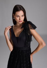 Robe noire avec un corsage ajusté et une jupe en tulle fluide. Présente un détail de nœud transparent sur une épaule et des motifs texturés sur le tissu.