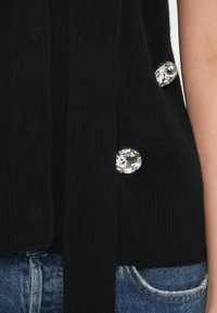 Top negro sin mangas de punto con acabado texturizado, que presenta grandes botones redondos brillantes en el lateral y un detalle de lazo en la cintura.