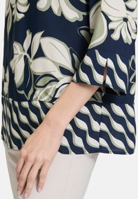 Granatowa bluzka w kwiaty z zielonymi i kremowymi wzorami. Posiada luźny krój, rękawy o długości 3/4 oraz rozcięty dół.