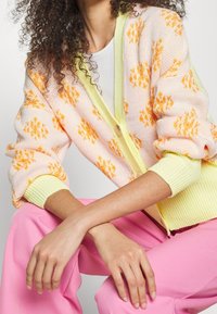 Vrouw met krullend haar, gekleed in een pastelgeel en oranje gebloemd vest over een wit shirt, gecombineerd met felroze broek, zittend met gekruiste armen.