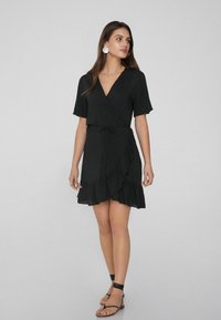 Schwarzes Wickelkleid mit kurzen Ärmeln, gerüschtem Saum und einem Bindegürtel. Hergestellt aus einem leichten Stoff, mit einem tiefen V-Ausschnitt.