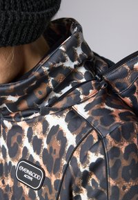 Treningsjakke med leopardmønster i brunt, svart og hvitt. Har høy krage og glidelåsdetaljer. Materialet ser glatt ut.