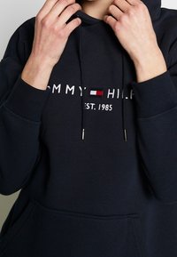 Sudadera azul marino con cordones, que presenta bordado blanco "TOMMY HILFIGER EST. 1985" y un logo con bloques de color. Incluye un bolsillo tipo canguro en el frente.