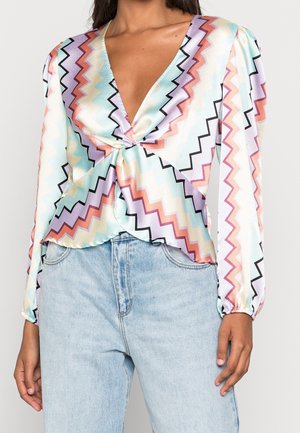 Blusa de manga larga con cuello en V y patrón de zigzag en tonos pastel de púrpura, naranja, azul y crema, combinada con vaqueros de tiro alto en azul claro.