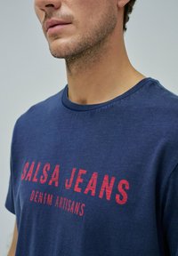 Camiseta de marina con el texto rojo "SALSA JEANS" y "DENIM ARTISANS" impreso en la parte frontal. Material de algodón suave; cuello redondo; mangas cortas.