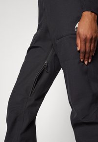 Black outdoor pants made from durable fabric, featuring a side zippered pocket and a smooth texture. Small logo in the upper area.