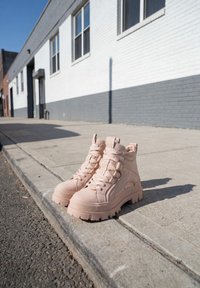 Paire de bottines épaisses rose clair à lacets posées sur un trottoir à côté d'un trottoir devant un immeuble en briques blanches et grises.