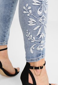 Höga ljusblå jeans med vit blommönstring på sidan, tillsammans med svarta klackar med ankelslinga och gyllene kedjedetaljer.