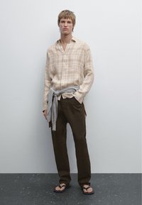 Camicia a quadri beige con bottoni, maglione grigio legato in vita, pantaloni marrone scuro e infradito neri. Sfondo a superficie opaca.