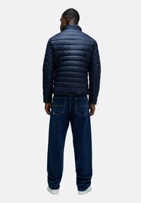 Granatowa kurtka puchowa z poziomym pikowaniem, w połączeniu z luźnymi niebieskimi dżinsami i białymi trampkami, widziana z tyłu.