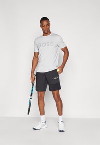 Grå t-skjorte med korte ermer og "BOSS"-logo, kombinert med svarte shorts med lomme. Hvite joggesko og et tennisracket i hånden.
