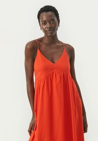 V-ringad ärmlös klänning i klar orange, med en strukturerad tyg, plisserad bodice och flödig kjol. Guld hoopsörhängen kompletterar looken.