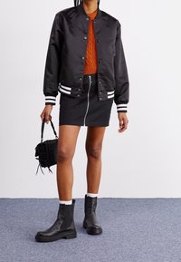 Schwarze Bomberjacke mit weißen Streifen, orangefarbener Kabelstrickpullover, schwarzer Reißverschlussrock und schwarze Stiefeletten mit weißen Akzenten. Schwarze Tasche.