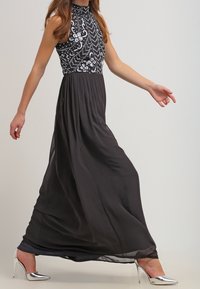 Schwarzes Abendkleid mit hohem, verzierten Ausschnitt und fließendem Rock. Es hat paillettenbesetzte Blumenmuster und silberne Stiletto-Absätze.