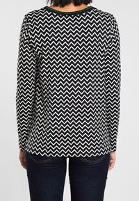 Černobílý top s dlouhým rukávem se zigzag vzorem, má kulatý výstřih a zelený akcent u límce, lem je ve zigzagovém stylu.