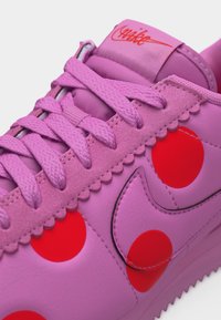 Μωβ αθλητικό παπούτσι Nike με ροζ κορδόνια, κόκκινα πουά, κυματιστές σουέντ λεπτομέρειες και κόκκινο λογότυπο Nike στη γλώσσα.