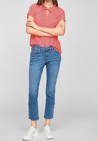 Blouse rouge à manches courtes avec un motif, dotée d'un col à nouer, associée à un jean bleu taille haute avec des boutons décoratifs à l'ourlet et des baskets blanches.