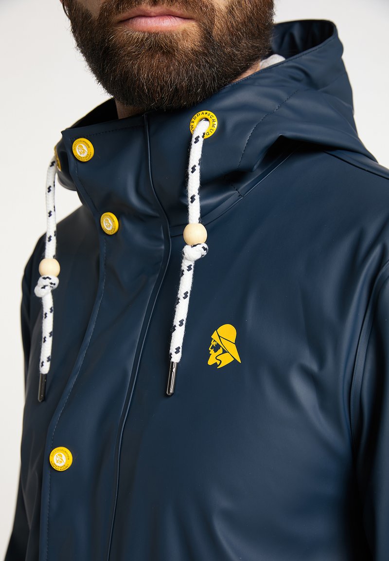 Marineblauer wasserdichter Jacke mit einem Frontreißverschluss, gelben Knöpfen und einem Logo. Zu den Merkmalen gehören eine Kapuze mit Kordelzügen und ein strukturiertes Gewebe.