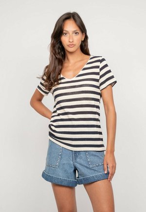 Femme portant un t-shirt à manches courtes à rayures noires et blanches avec un col en V et un short en denim retroussé avec des poches cloutées, se tenant devant un fond uni.