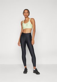 Sujetador deportivo verde claro con tirantes gruesos, combinado con leggings negros de cintura alta. Textura suave, diseño ajustado, con un branding mínimo en los leggings.