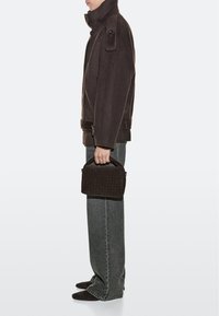 Personne portant un manteau marron foncé et un jean gris, tenant un petit sac à main tissé marron foncé, debout de profil devant un fond uni.