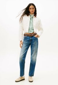 Chaqueta blanca corta con cuello marrón, camiseta a rayas verdes y blancas, jeans rectos azules y zapatillas beige con suelas oscuras.