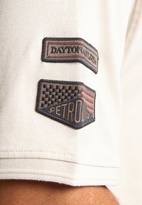 Polo shirt van licht crèmekleurige stof met twee textuur patches: één met "DAYTON" en "NATIONAL" in donkerrood, de ander met een geblokt patroon en "PETRON."