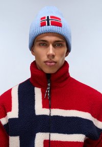 Jachetă din fleece roșie și bleumarin cu un model inspirat din steagul norvegian; asociată cu o căciulă tricotată de culoare bleu deschis cu broderie a steagului. Detaliu cu guler cu fermoar.