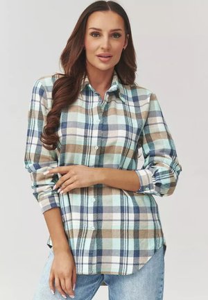 Kobieta z długimi brązowymi włosami nosi niebiesko-beżowo-białą koszulę w kratę zapinaną na guziki, z podwiniętymi rękawami, oraz jasnoniebieskie dżinsy na jednolitym tle.