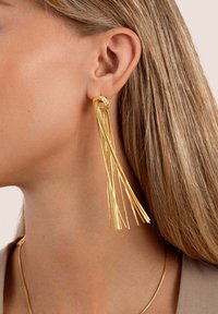 Orecchini in tono oro caratterizzati da un dettaglio a nodo con più fili che scendono, che mostrano una texture metallica lucida e un design elegante.