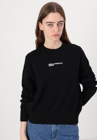 Črna pletena kapuca s krožnim izrezom, ribastimi manžetami in pasom, z belim vezenim logotipom "KARL LAGERFELD JEANS" spredaj.