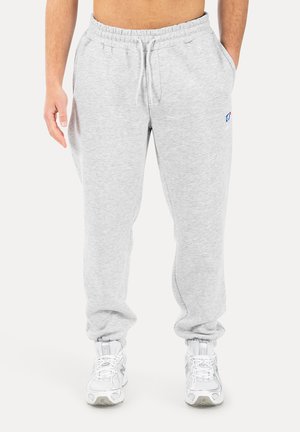 Uomo che indossa pantaloni da ginnastica grigio chiaro con vita elasticizzata e coulisse, abbinati a scarpe sportive bianche, in piedi contro uno sfondo bianco.