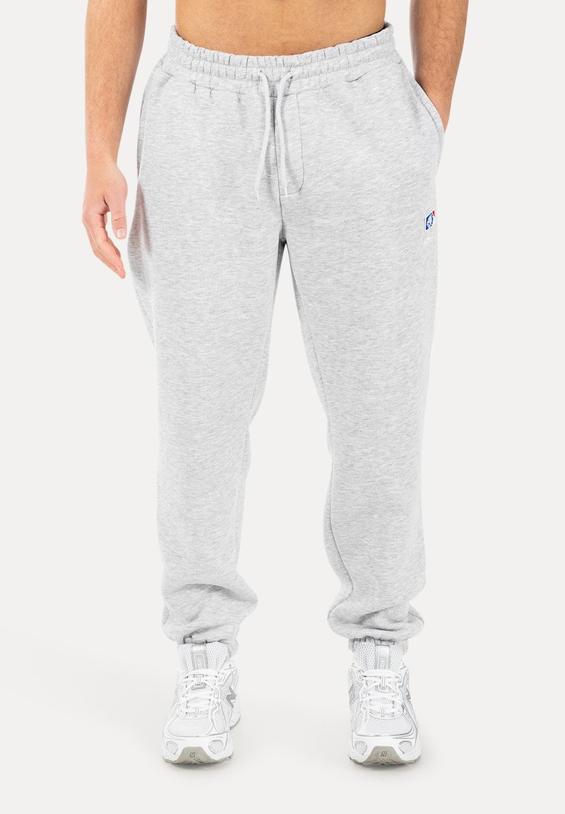 Mann trägt hellgraue Jogginghose mit elastischem Bund und Kordelzug, kombiniert mit weißen Sportschuhen, steht vor weißem Hintergrund.